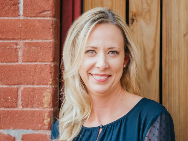Blonde-haired woman wearing a dark blue shirt, smiling and standing next to a brick wall